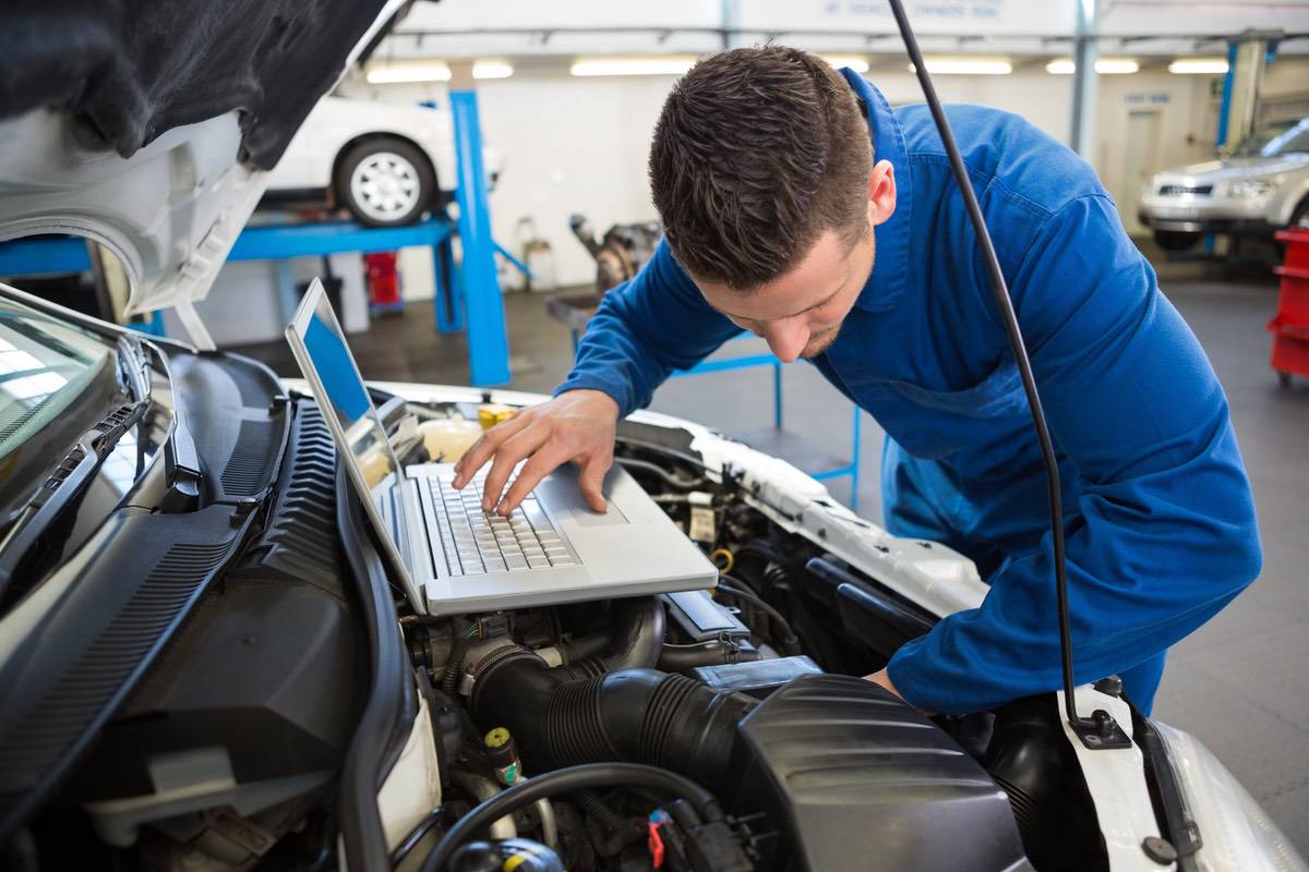 A mechanical engineer stands over a car engine with a laptop resting on the engine.