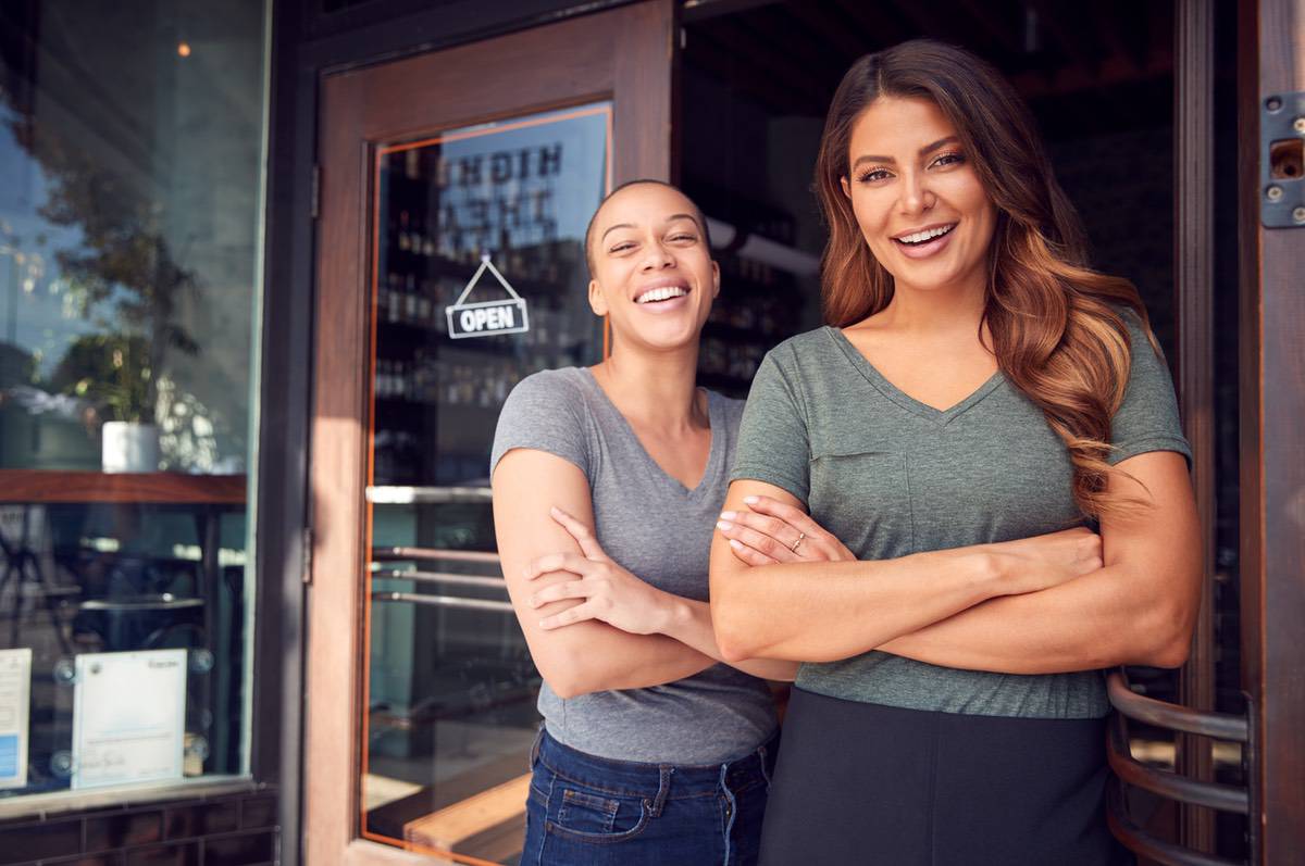 Two women stand in front of a store arms crossed smiling at the camera