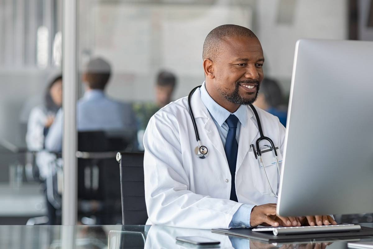 A man with a white coat and stethoscope around his neck smiles at a computer screen.